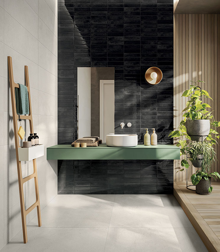 Bagno contemporaneo con rivestimenti delle collezioni La Matière Talc e Workshop Core, caratterizzato da una parete scura lucida, superfici chiare, mobile lavabo verde sospeso, dettagli in legno e numerose piante illuminate dalla luce naturale.