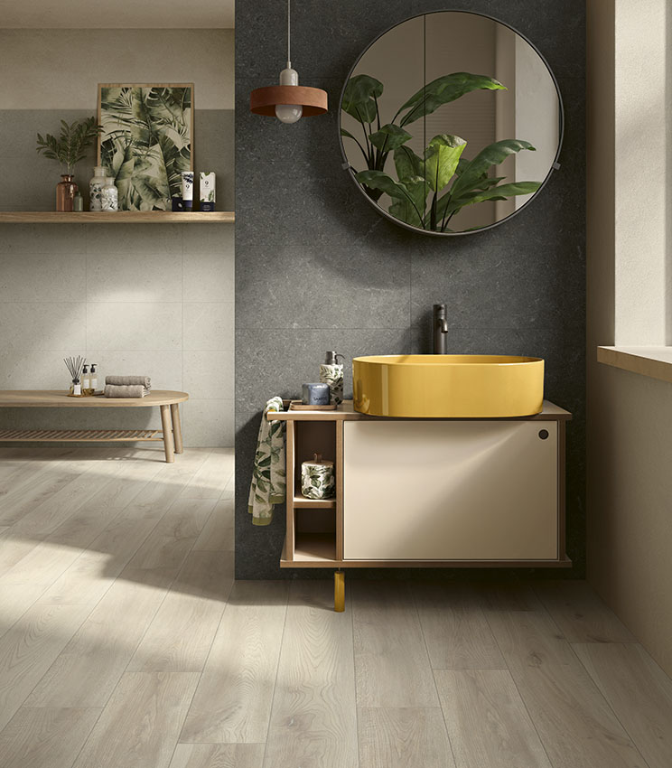 Bagno contemporaneo con superfici delle collezioni Surround Outline Soft, Draft e Borealis Inari, caratterizzato da una parete scura, lavabo ovale giallo, mobile sospeso, grande specchio rotondo, piante decorative e pavimento effetto legno chiaro.