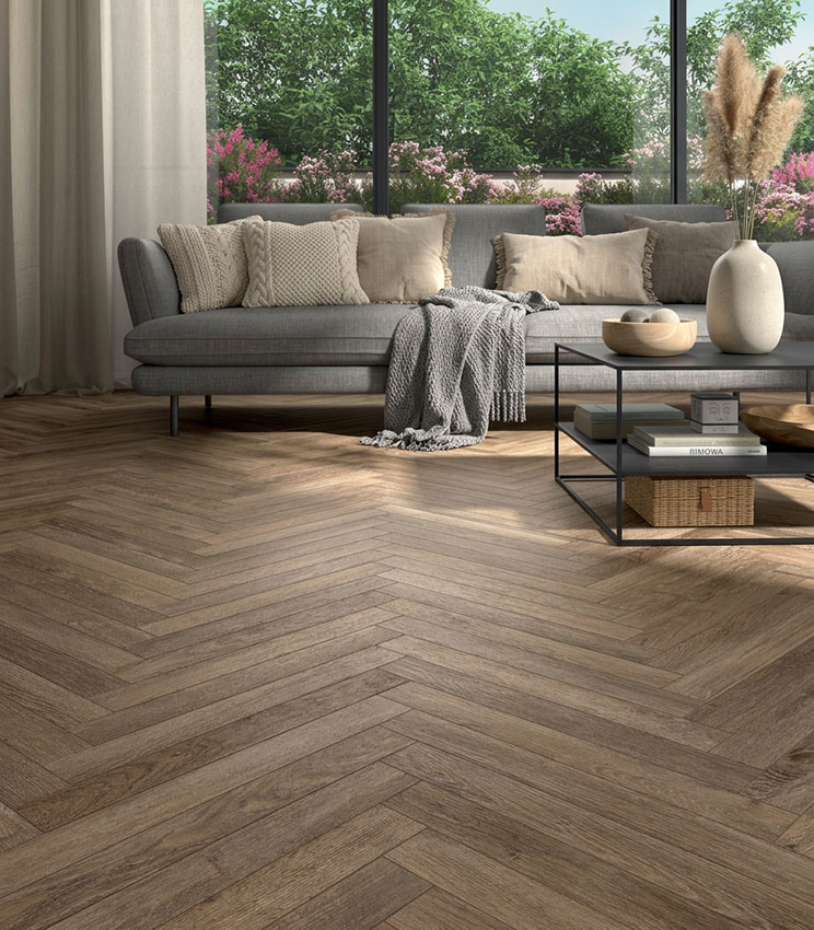 Soggiorno contemporaneo con pavimento della collezione Kairos Stigmi posato a spina di pesce, arredato con ampio divano grigio, tavolino in metallo, tessili dai toni neutri e grandi vetrate affacciate su un giardino fiorito.