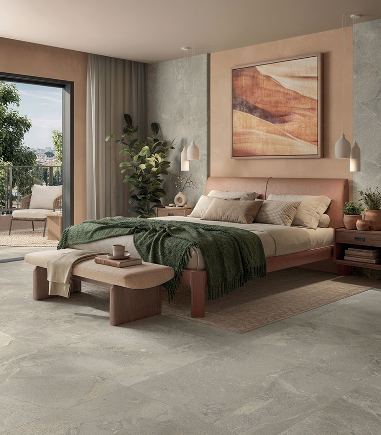 Camera da letto contemporanea con superfici della collezione Revel Ground, caratterizzata da pavimento e rivestimenti effetto pietra, letto imbottito nei toni rosa cipria, tessili neutri e verde scuro, comodini in legno e ampia apertura verso una terrazza luminosa.