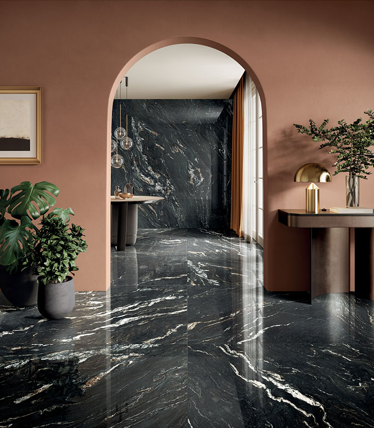 Ambiente contemporaneo con superfici della collezione Perpetual Belvedere Dark Lux, caratterizzato da grandi lastre scure effetto marmo con venature chiare e finitura lucida. Lo spazio è completato da pareti color terracotta, arredi dalle linee essenziali, piante decorative, lampade dorate e un’apertura ad arco.