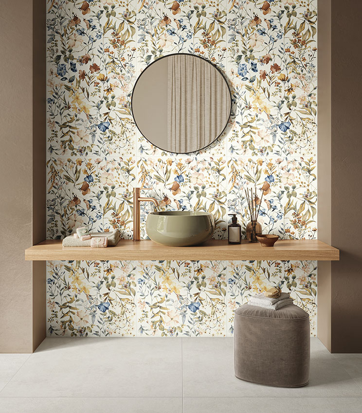 Bagno contemporaneo con superfici delle collezioni La Matière Talc e La Fleur, caratterizzato da un rivestimento floreale nei toni naturali, lavabo verde oliva, mensola in legno, specchio rotondo e pouf imbottito su pavimento chiaro.