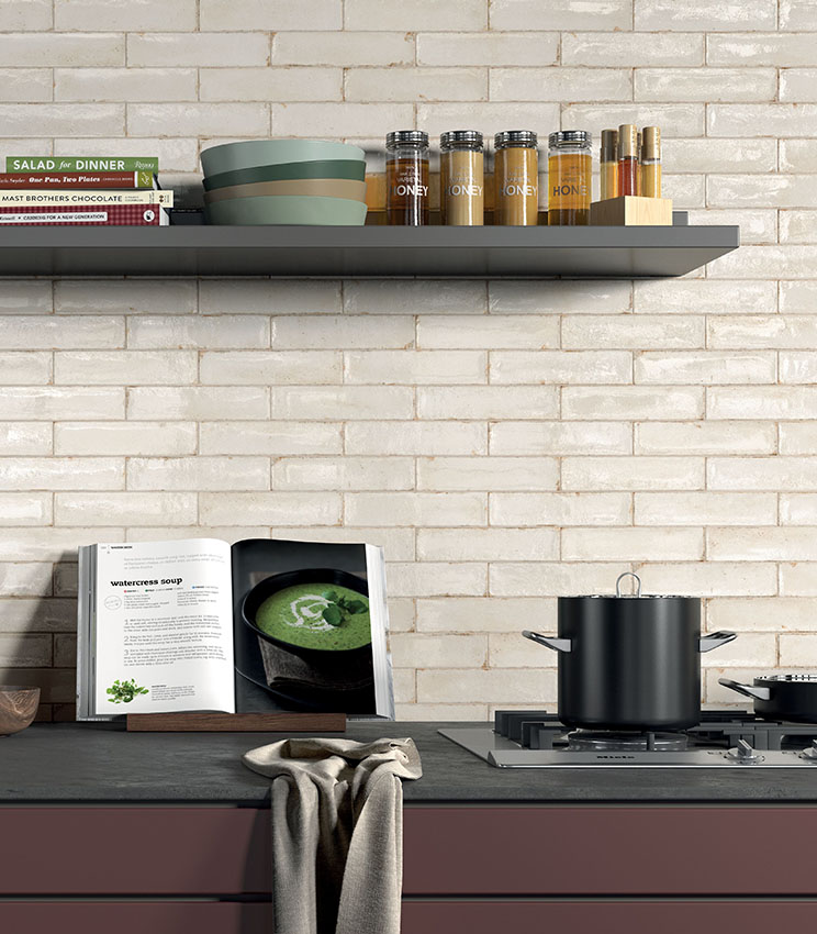 Dettaglio di cucina contemporanea con rivestimento della collezione Workshop Opal, caratterizzato da piastrelle rettangolari chiare con finitura irregolare e lucida. L’ambiente presenta mensole scure, piano di lavoro nero, mobili color bordeaux, libri di cucina, contenitori e pentole.