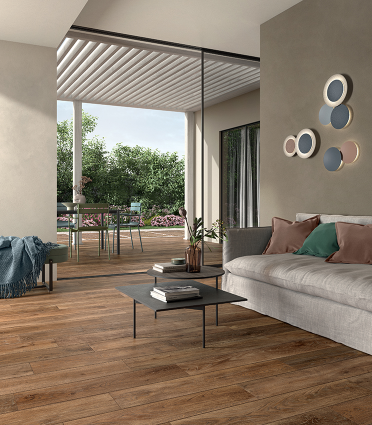 Soggiorno contemporaneo con pavimento della collezione Kairos Zoi, caratterizzato da superfici effetto legno dai toni caldi, ampio divano chiaro, tavolini bassi e accesso diretto a una terrazza arredata affacciata sul verde.