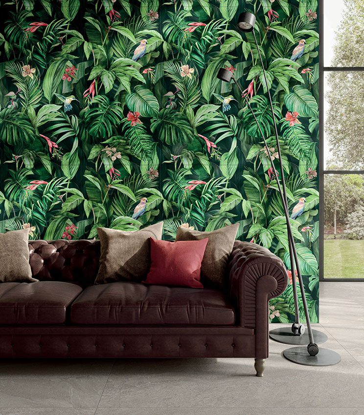 Soggiorno contemporaneo con rivestimento decorativo della collezione Glam Vision, caratterizzato da una parete con motivo tropicale di foglie, fiori e uccelli, divano in pelle scura con cuscini nei toni neutri e bordeaux, lampade da terra nere e ampia vetrata affacciata sul giardino.