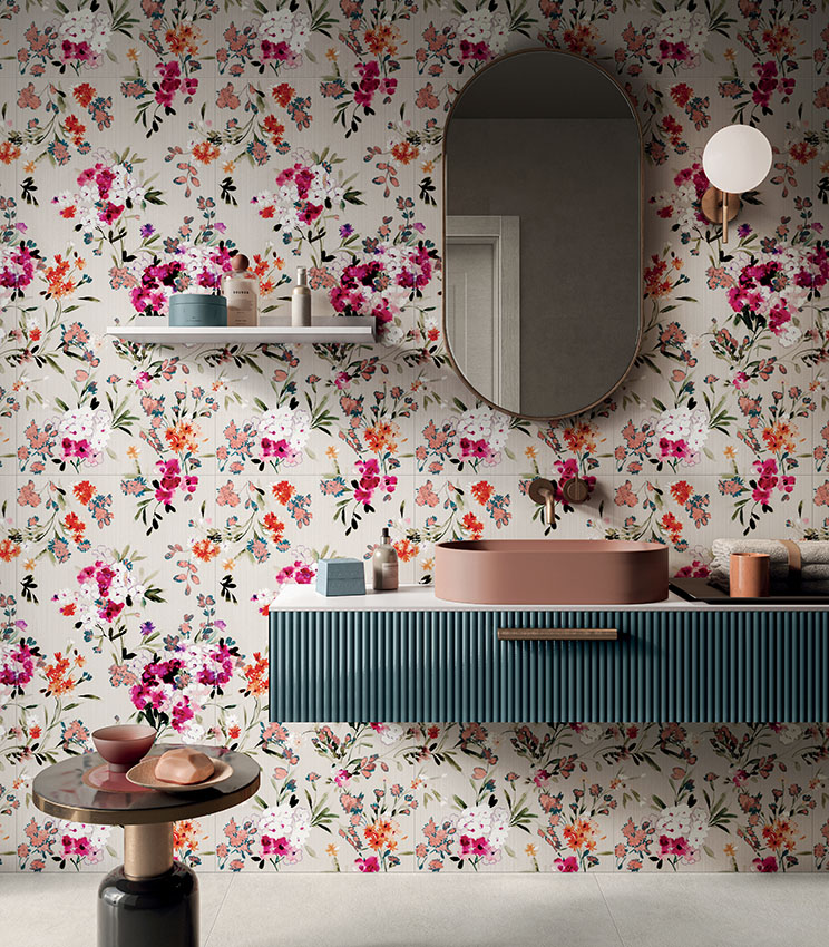Bagno contemporaneo con rivestimento decorativo della collezione Glam Bouquet, caratterizzato da un motivo floreale multicolore su fondo chiaro, mobile lavabo sospeso blu petrolio, lavabo rosa cipria, grande specchio ovale e dettagli coordinati nei toni caldi.