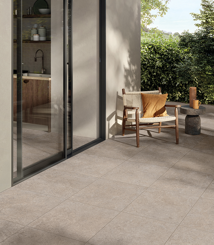 Terrazza contemporanea con pavimentazione della collezione Surround Row, caratterizzata da grandi lastre effetto pietra nei toni beige, poltrona in legno e corda, tavolino scultoreo e vegetazione rigogliosa sullo sfondo. Le ampie vetrate collegano lo spazio esterno alla cucina interna.