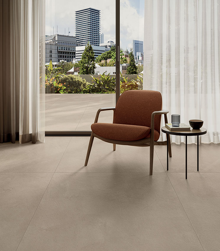Scorcio di living moderno, con poltrona e porta scorrevole che dà su un terrazzo da cui si gode una vista metropolitana. Pavimento effetto cemento di Panaria Ceramica, collezione La Matière.