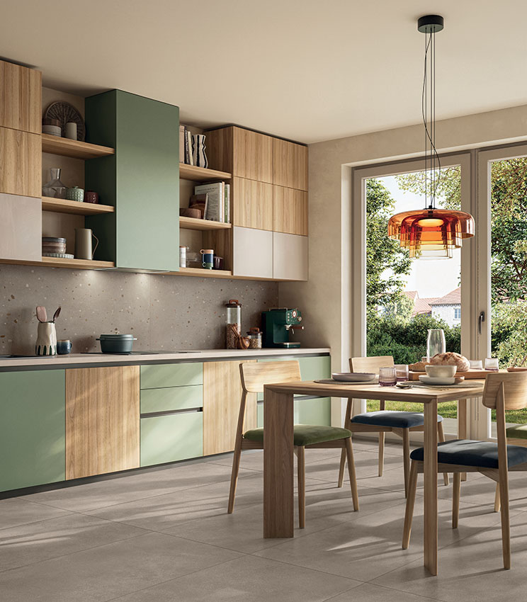 Cucina moderna e accogliente con arredi in legno e verde salvia, zona pranzo luminosa e pavimento in gres porcellanato effetto cemento dai toni caldi.