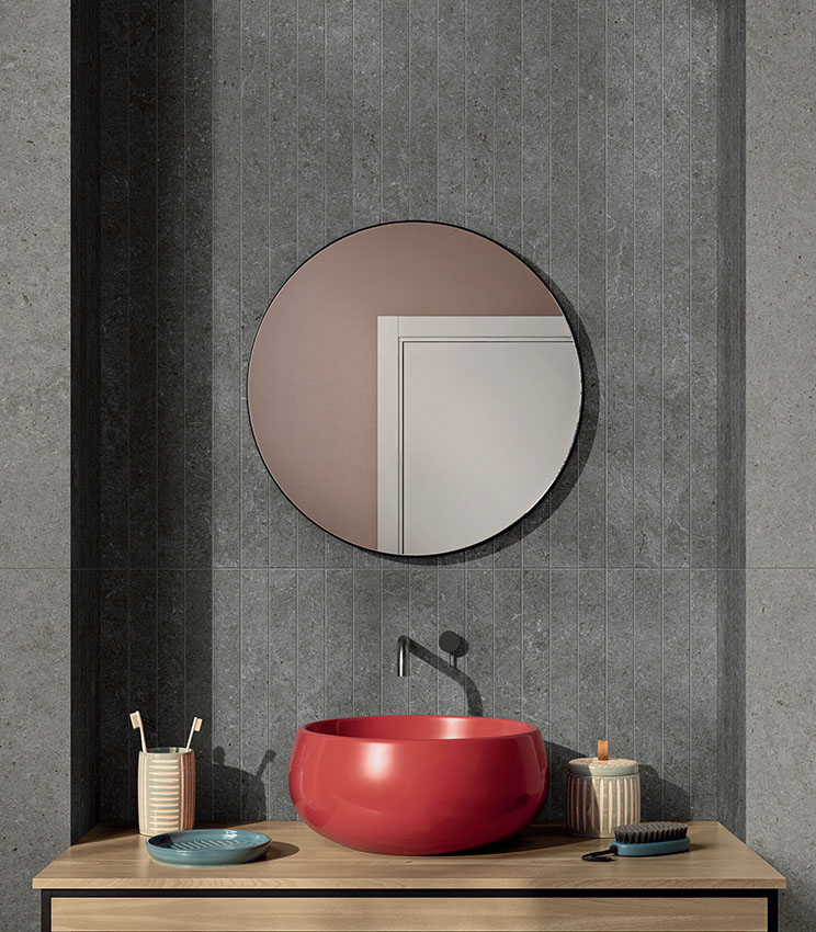 Bagno rivestito con piastrelle di gres porcellanato effetto pietra, doccial walk-in, specchio tondo e lavabo esterno rosso.