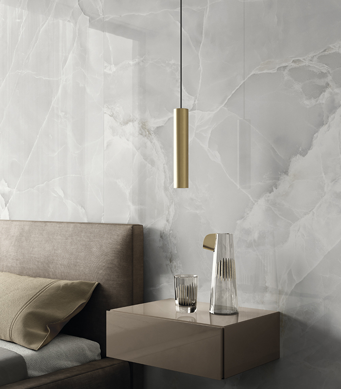 Dettaglio di parete rivestita con gres porcellanato effetto marmo chiaro in finitura lucida, con comodino sospeso, punto luce sospeso e scorcio di letto matrimoniale.