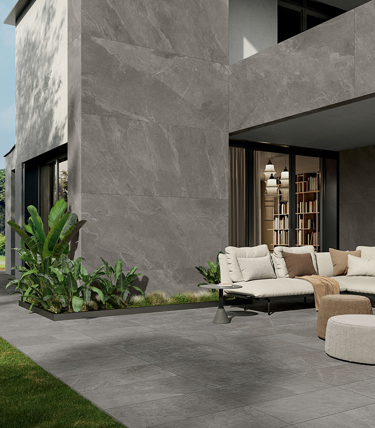 Veduta esterna di villa dal design moderno e rigorosamente geometrico, parzialmente rivestita con grandi lastre ceramiche effetto pietra, con patio arredato pavimentato con gres porcellanato effetto pietra in 20 mm di spessore.