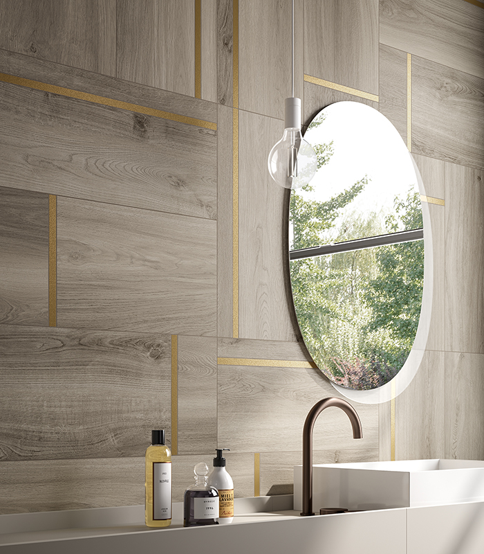 Dettaglio della parete lavabo di un bagno moderno, con lavabo lineare, specchio ovale e rivestimento in gres porcellanato decorativo effetto legno color cenere con inserti ottone.