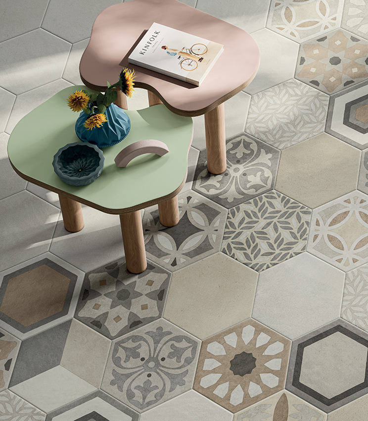 Dettaglio di pavimento con superfici delle collezioni La Matière Hexa Talc Sable e Hexa Chic, caratterizzato da piastrelle esagonali in tonalità neutre, alternate a motivi decorativi geometrici e floreali. Due tavolini dalle forme morbide, in verde e rosa, completano la scena con vasi e piccoli oggetti decorativi.
