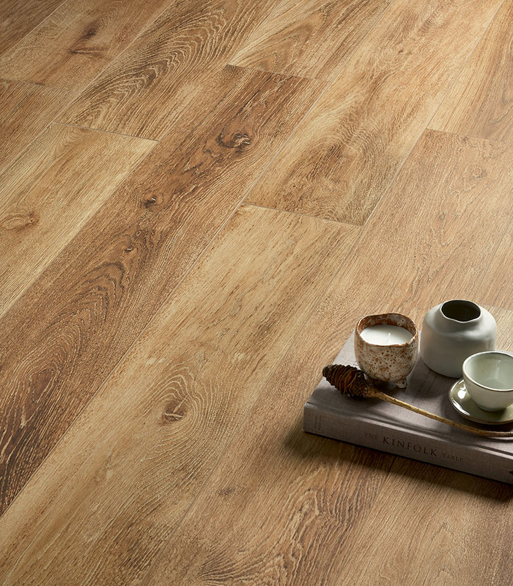 Dettaglio di pavimento della collezione Kairos Ilios, caratterizzato da superfici effetto legno con venature marcate e tonalità calde e naturali. La scena è completata da un libro, una candela, un piccolo vaso e una tazza in ceramica.