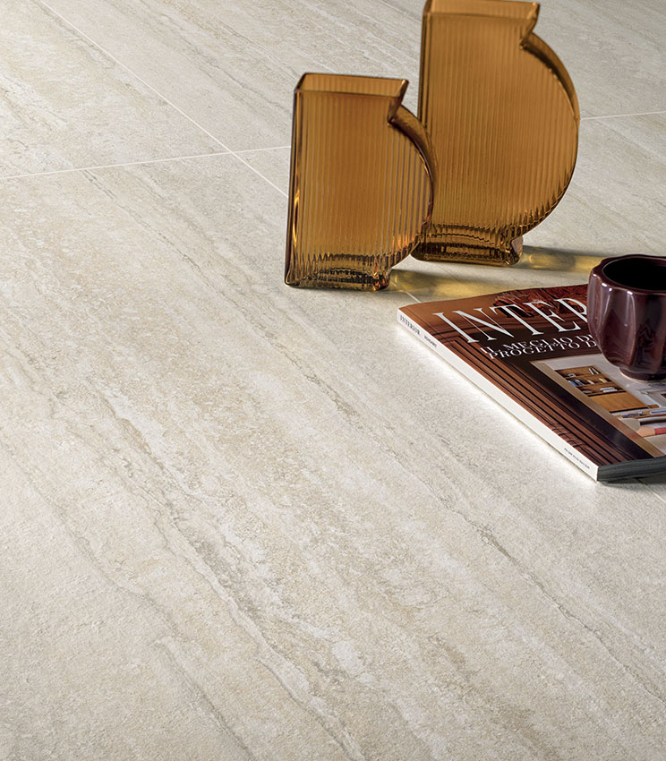 Dettaglio di pavimento della collezione Perpetual Travertino Ivory, caratterizzato da superfici effetto travertino nei toni chiari del beige e dell’avorio. La scena è completata da due vasi in vetro ambrato, una rivista e una tazza scura.
