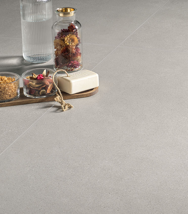 Dettaglio di pavimento della collezione Formula Form Ash, caratterizzato da una superficie chiara e uniforme dall’aspetto materico. La scena è completata da una composizione con bottiglia in vetro, vaso con fiori secchi, piccoli contenitori e una saponetta su vassoio in legno.