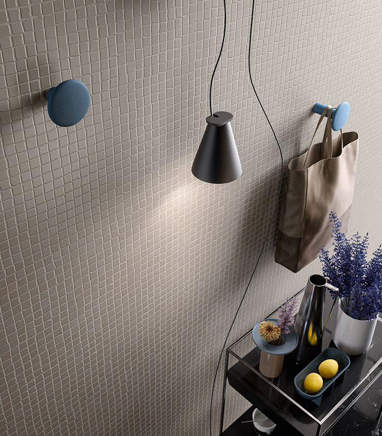 Dettaglio di rivestimento della collezione Even Dove Mosaico, caratterizzato da una trama regolare di piccole tessere quadrate nei toni chiari. La scena è completata da ganci blu, lampada a sospensione nera, borsa in tessuto e una consolle con vasi, fiori e piccoli oggetti decorativi.
