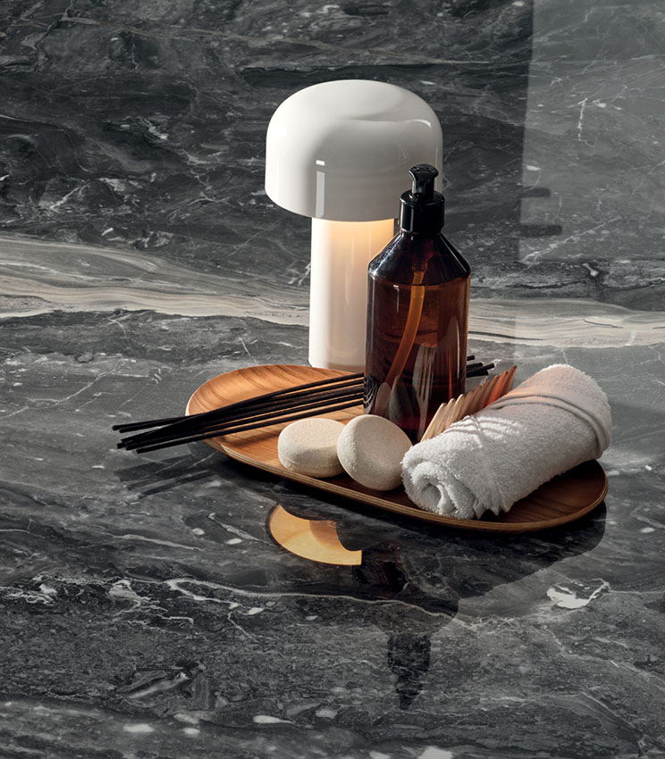 Dettaglio di superficie della collezione Perpetual Orobico Blend Lux, caratterizzata da un effetto marmo scuro con venature chiare e finitura lucida. La scena è completata da un vassoio in legno con flacone, asciugamano arrotolato, piccoli accessori e una lampada da tavolo bianca.