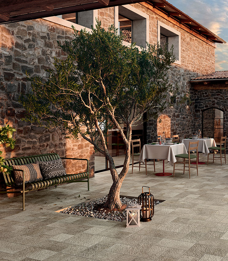 Terrazza di ristorante con pavimentazione della collezione Pierre des Rêves Aurore, caratterizzata da superfici effetto pietra in tonalità naturali. L’ambiente, circondato da pareti in pietra, è arredato con tavoli e sedie in legno, una panca, lanterne decorative e un ulivo al centro dello spazio.