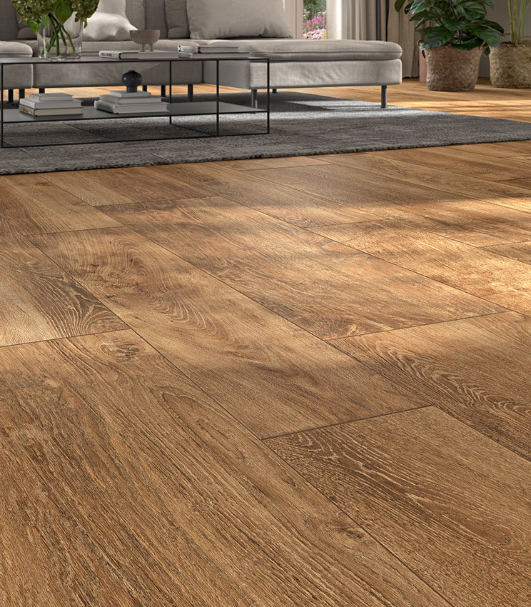 Soggiorno contemporaneo con pavimento della collezione Kairos Ilios, caratterizzato da superfici effetto legno in tonalità calde e da venature marcate. L’ambiente è arredato con ampio divano grigio, tavolino in metallo nero, tappeto scuro e piante decorative illuminate dalla luce naturale.