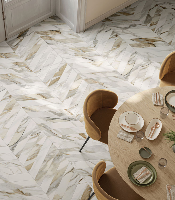 Sala da pranzo contemporanea con pavimento della collezione Perpetual Venato Gold Soft Chevron, caratterizzato da un motivo a chevron effetto marmo bianco con venature grigie e dorate. L’ambiente è arredato con tavolo rotondo in legno chiaro, sedie imbottite color ocra e mise en place dai toni neutri.