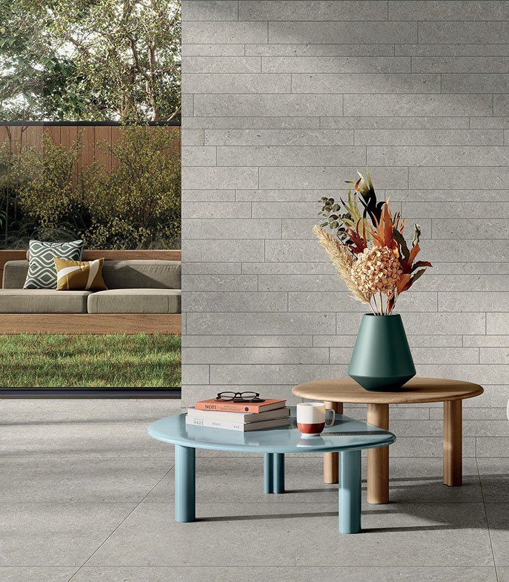Soggiorno contemporaneo con superfici delle collezioni Surround Shape e Shape Muretto Soft, caratterizzato da pavimento effetto pietra grigia e parete rivestita con elementi rettangolari coordinati. L’ambiente è arredato con tavolini dalle linee essenziali, vaso con composizione floreale e ampia apertura verso un giardino con divano esterno.