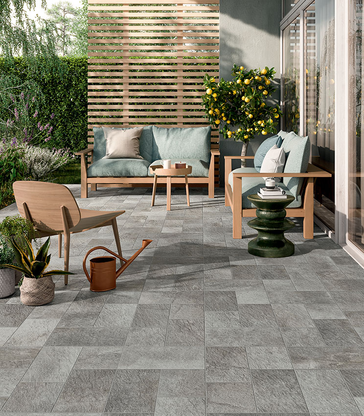 Terrazza contemporanea con pavimentazione della collezione Pierre des Rêves Nuage, caratterizzata da superfici effetto pietra grigia in formati modulari. L’ambiente è arredato con divani e poltrone in legno, cuscini verde salvia, tavolini decorativi, piante in vaso e un agrume, immersi in un giardino rigoglioso.