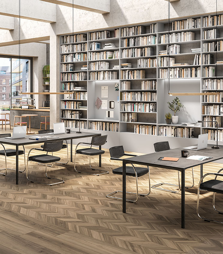 Biblioteca e spazio di lavoro contemporaneo con pavimento della collezione Nuance Cendre e Nuance Cendre Chevron, caratterizzato da superfici effetto legno in tonalità naturali e posa geometrica. L’ambiente presenta grandi librerie a parete, tavoli da lavoro con sedie nere, laptop, piante decorative e ampie finestre che illuminano lo spazio.