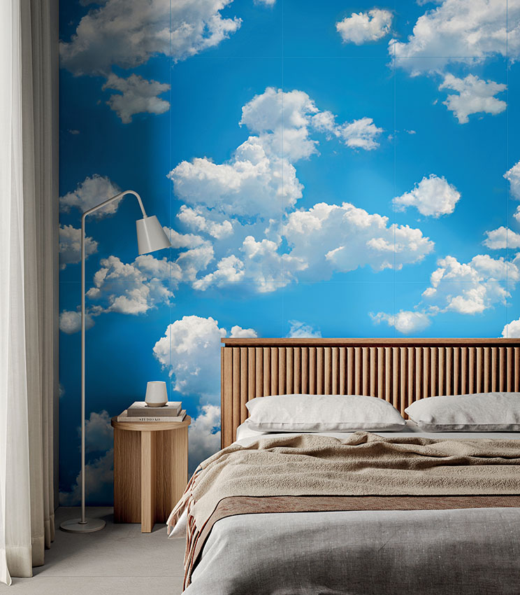 Camera da letto contemporanea con rivestimento decorativo della collezione Glam Flow, caratterizzato da un cielo azzurro con grandi nuvole bianche. L’ambiente è arredato con letto dalla testiera in legno scanalato, biancheria dai toni neutri, comodino in legno e lampada da terra bianca.
