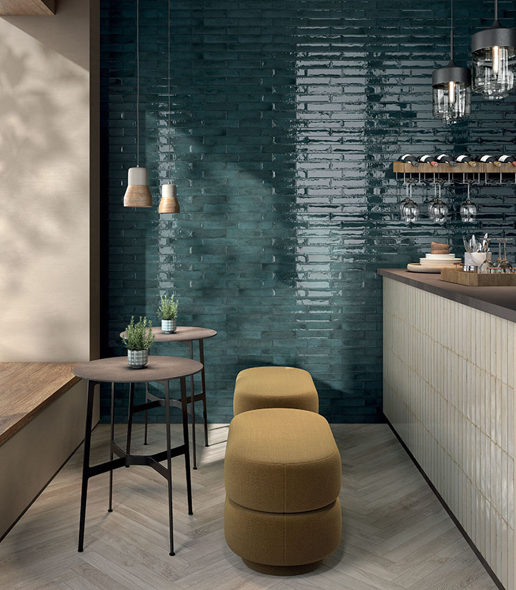 Angolo bar contemporaneo con superfici delle collezioni Workshop Berry, Workshop Opal e Kairos Imera, caratterizzato da una parete verde petrolio lucida, bancone rivestito in tonalità chiare, pavimento effetto legno e arredi compatti con sgabelli imbottiti e tavolini alti.