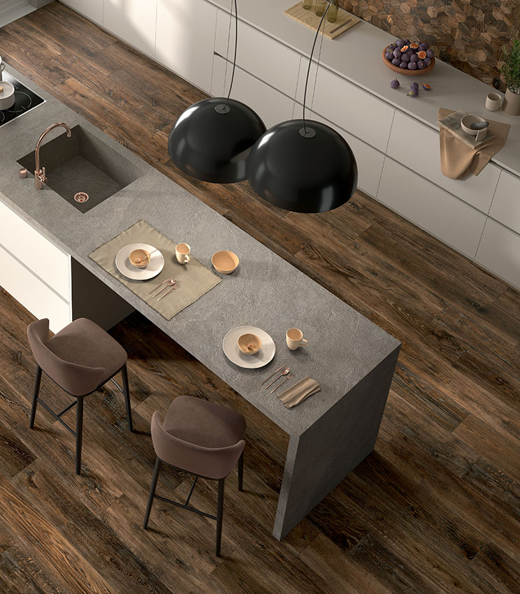 Vista dall'alto di una cucina moderna con isola dinette rivestita con lastre di gres effetto pietra color grigio chiaro e con pavimento in doghe scure di gres porcellanato effetto legno