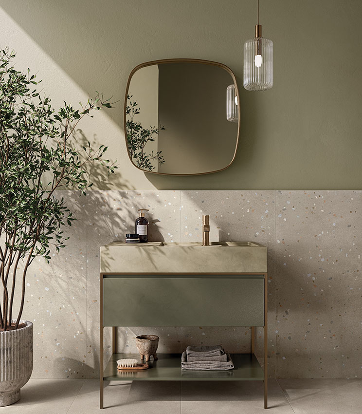 Bagno contemporaneo con superfici delle collezioni Formula Form Rope e Seed Rope, caratterizzato da un rivestimento chiaro con piccoli inserti colorati, pareti verde salvia, mobile lavabo coordinato, grande specchio sagomato, lampada a sospensione e pianta decorativa.