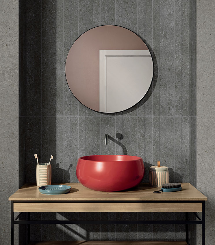 Bagno contemporaneo con superfici delle collezioni Surround Shape Soft e Outline Carving, caratterizzato da rivestimenti grigi effetto pietra e da una fascia centrale con motivo lineare verticale. L’ambiente presenta un lavabo rosso lucido su piano in legno, specchio rotondo, rubinetteria scura e piccoli accessori da bagno.