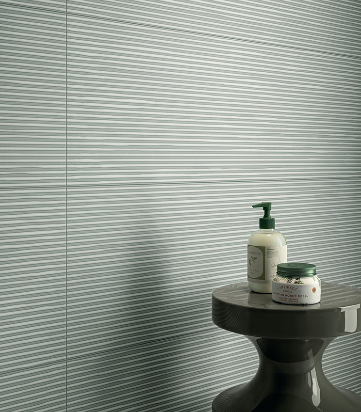 Dettaglio di bagno contemporaneo con rivestimento della collezione Lullaby Linear Green, caratterizzato da una superficie verde chiaro con motivo lineare orizzontale. La scena è completata da un tavolino scuro dalla forma scultorea con flaconi e piccoli prodotti da bagno.