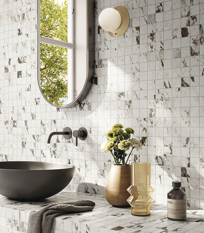 Scorcio della parete lavabo di un bagno piccolo rivestito con mosaico ceramico effetto marmo chiaro, con vasca lavabo scura in appoggio e specchio ovale