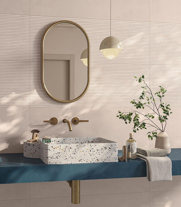 Bagno contemporaneo con superfici delle collezioni Lullaby Fairytale Blush e Linear Blush, caratterizzato da rivestimenti rosa cipria con fasce a rilievo lineare. L’ambiente presenta lavabo rettangolare effetto terrazzo, piano blu, specchio ovale con cornice dorata, rubinetteria coordinata, lampada a sospensione e rami verdi decorativi.