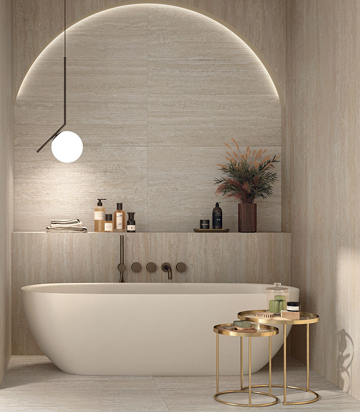 Bagno contemporaneo con superfici delle collezioni Perpetual Travertino Ivory Edge, Travertino Ivory e Travertino Cream, caratterizzato da rivestimenti effetto travertino in tonalità calde e naturali. L’ambiente presenta una vasca freestanding bianca inserita in una nicchia ad arco retroilluminata, mensole integrate, lampada a sospensione e tavolini metallici dorati con accessori da bagno.