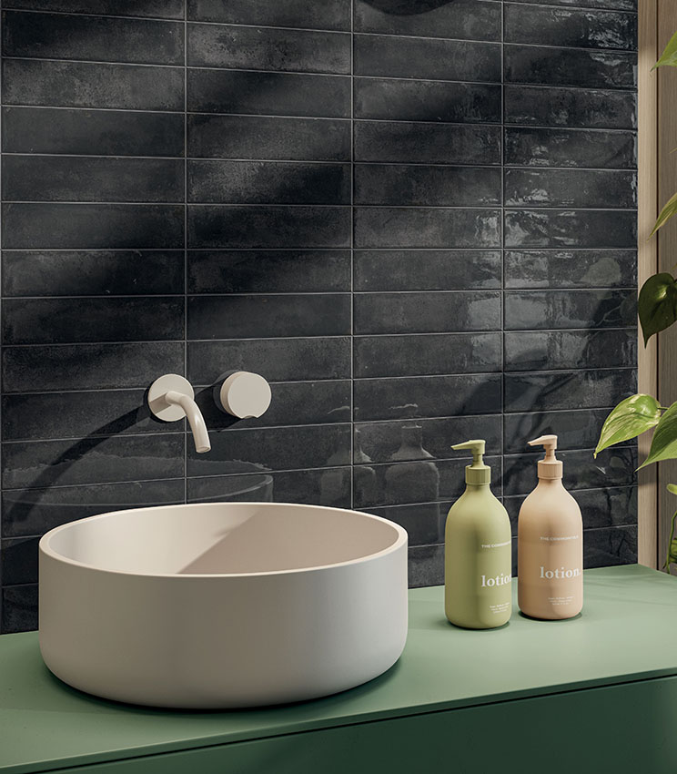 Dettaglio di bagno contemporaneo con rivestimento della collezione Workshop Core, caratterizzato da piastrelle rettangolari scure con finitura lucida e irregolare. La scena presenta un lavabo da appoggio bianco su mobile verde, rubinetteria a parete, flaconi coordinati e una pianta decorativa.