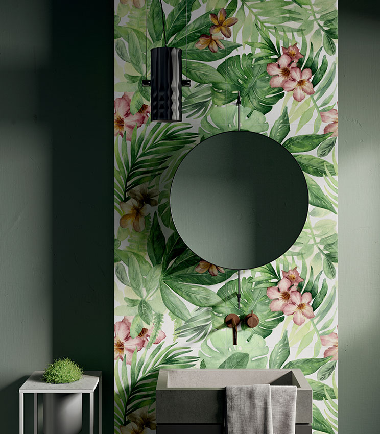 Bagno contemporaneo con rivestimento decorativo della collezione Glam Rainforest, caratterizzato da un motivo tropicale con grandi foglie verdi e fiori rosa. L’ambiente presenta pareti verde scuro, lavabo dalle linee squadrate, specchio rotondo, rubinetteria a parete e lampada a sospensione nera.