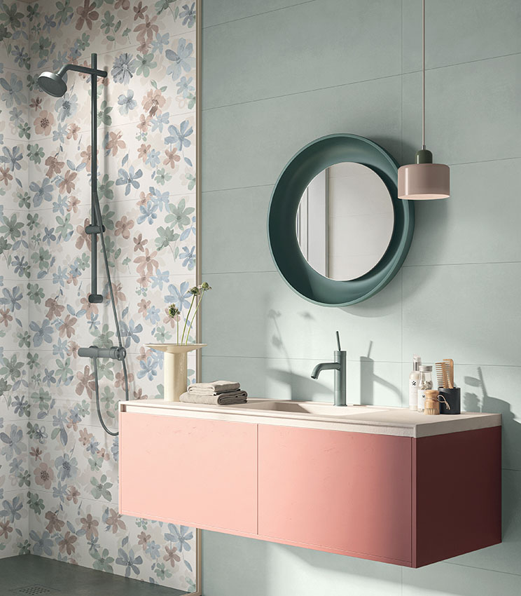 Bagno contemporaneo con superfici delle collezioni Lullaby Silent Green, Florè e Borealis Abisko, caratterizzato da rivestimenti verde chiaro e da una parete decorata con motivo floreale nei toni pastello. L’ambiente presenta mobile lavabo sospeso rosa, specchio rotondo verde scuro, rubinetteria coordinata, lampada a sospensione e zona doccia con colonna nera.