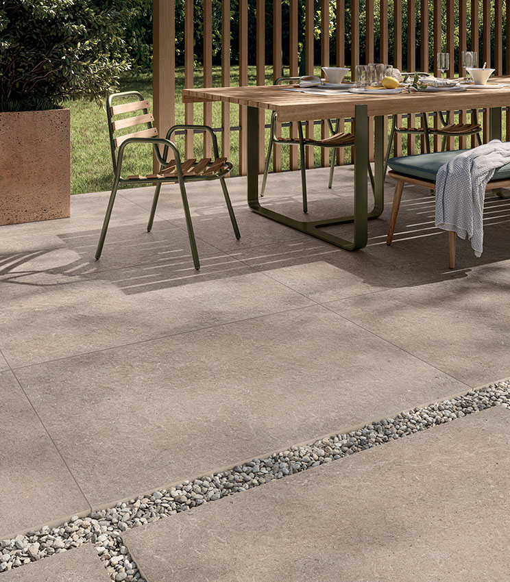 Scorcio di patio allestito con tavolo da colazione pavimentato con gres outdoor in 20 mm di spessore effetto pietra color sabbia. Il gres 20 mm è in parte posato a colla e in parte posato a secco su ghiaia. | Collezione Surround di Panaria Ceramica