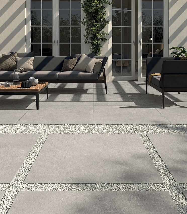 Scorcio di patio arredato con moderno salottino composto da divano e poltrona. Pavimento in gres porcellanato 20 mm effetto cemento, posato a colla. In primo piano, camminamento outdoor realizzato con grandi lastre ceramiche posate a secco su ghiaia. | Collezione Formula di Panaria Ceramica