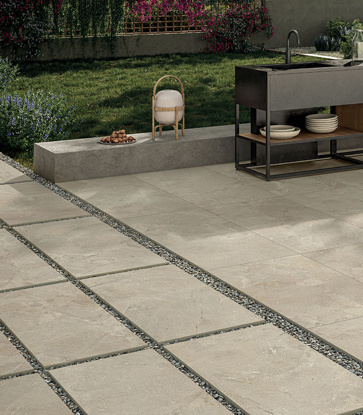 Scorcio di bordo piscina con moderno blocco cucina da esterni e giardino verde retrostante. Il pavimento, in gres porcellanato effetto pietra color sabbia, è posato a colla attorno alla piscina e, in primo piano, posato a secco su ghiaia a formare un camminamento. | Collezione Revel di Panaria Ceramica
