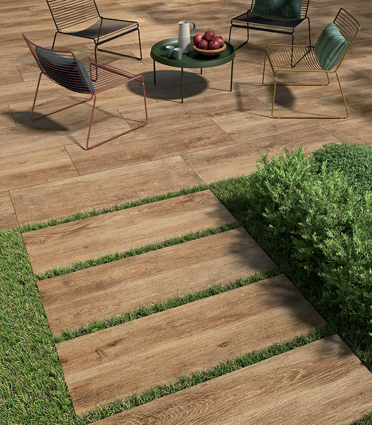 Scorcio di patio con moderne sedute di filo metallico e pavimento in gres porcellanato 20 mm effetto legno posato a colla. In primo piano parte un camminamento di doghe ceramiche outdoor posate a secco su erba. | Collezione Kairos di Panaria Ceramica