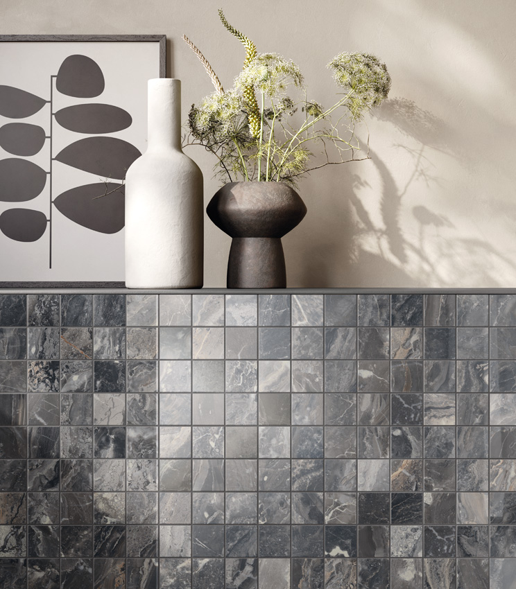 Vista ravvicinata e frontale di muretto con mensola su cui poggiano due vasi e un quadro astratto. Il muretto è rivestito di mosaico ceramico effetto marmo scuro mentre il resto della parete è tinteggiato color avorio.