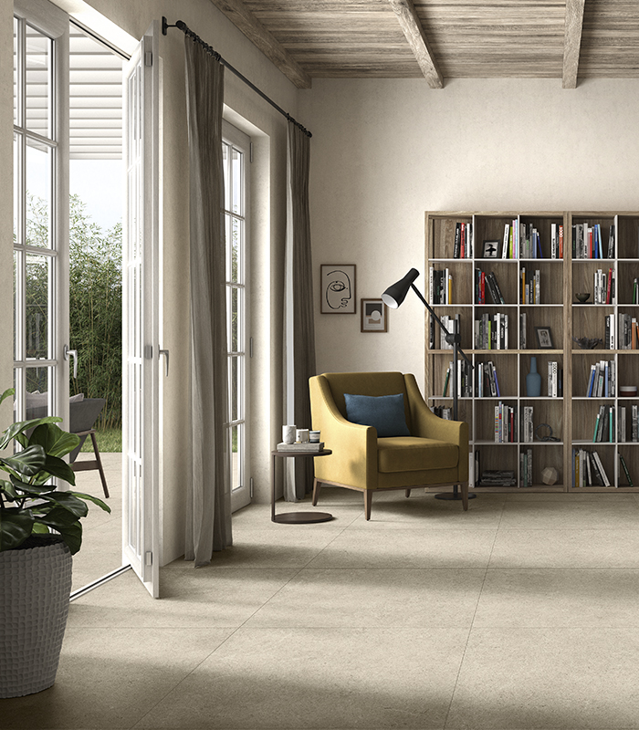 Accogliente living in stile rustico contemporaneo con grande libreria, poltrona da lettura giallo ocra e divano carta da zucchero, soffitto in travi di legno e due porte-finestre affacciate su un portico. Pavimento in gres porcellanato effetto pietra color sabbia.