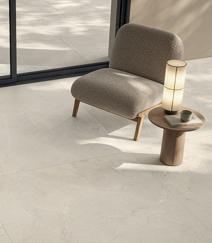 Living moderno e luminoso con poltrona imbottita, tavolino in legno e pavimento in gres porcellanato effetto pietra chiara, in continuità con lo spazio esterno.