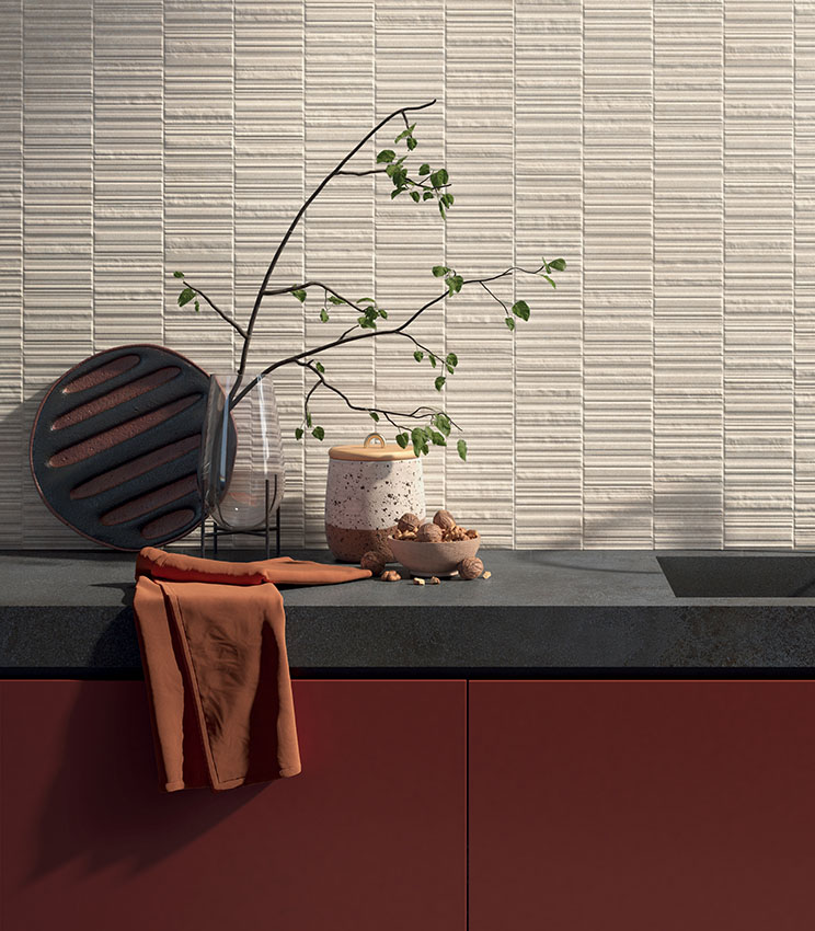 Cucina contemporanea con rivestimento della collezione Iqoniqa Brix Skin, caratterizzata da parete chiara a rilievo, piano di lavoro scuro, mobili color terracotta e composizione decorativa con vasi, rami verdi e oggetti in ceramica.