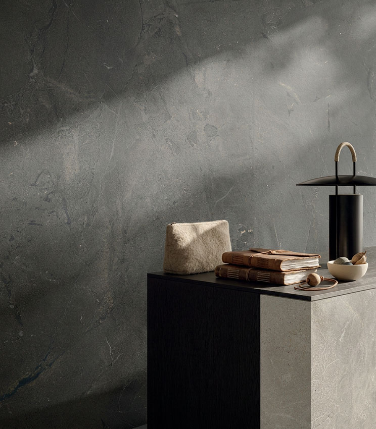 Dettaglio di superfici della collezione Revel Night e Revel Ground, con grandi lastre effetto pietra nei toni grigio scuro e beige. La scena è completata da una consolle minimale, libri, piccoli oggetti decorativi e una lampada nera, illuminati da luce naturale radente.